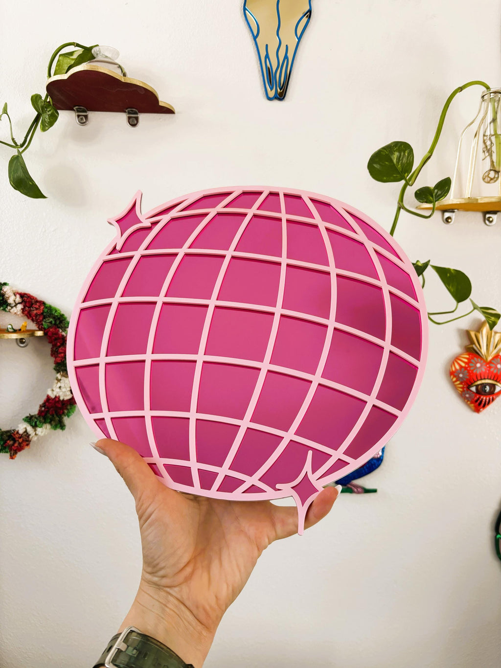 Hand holding a pink spherical object with grid pattern against a white wall with decorative items.