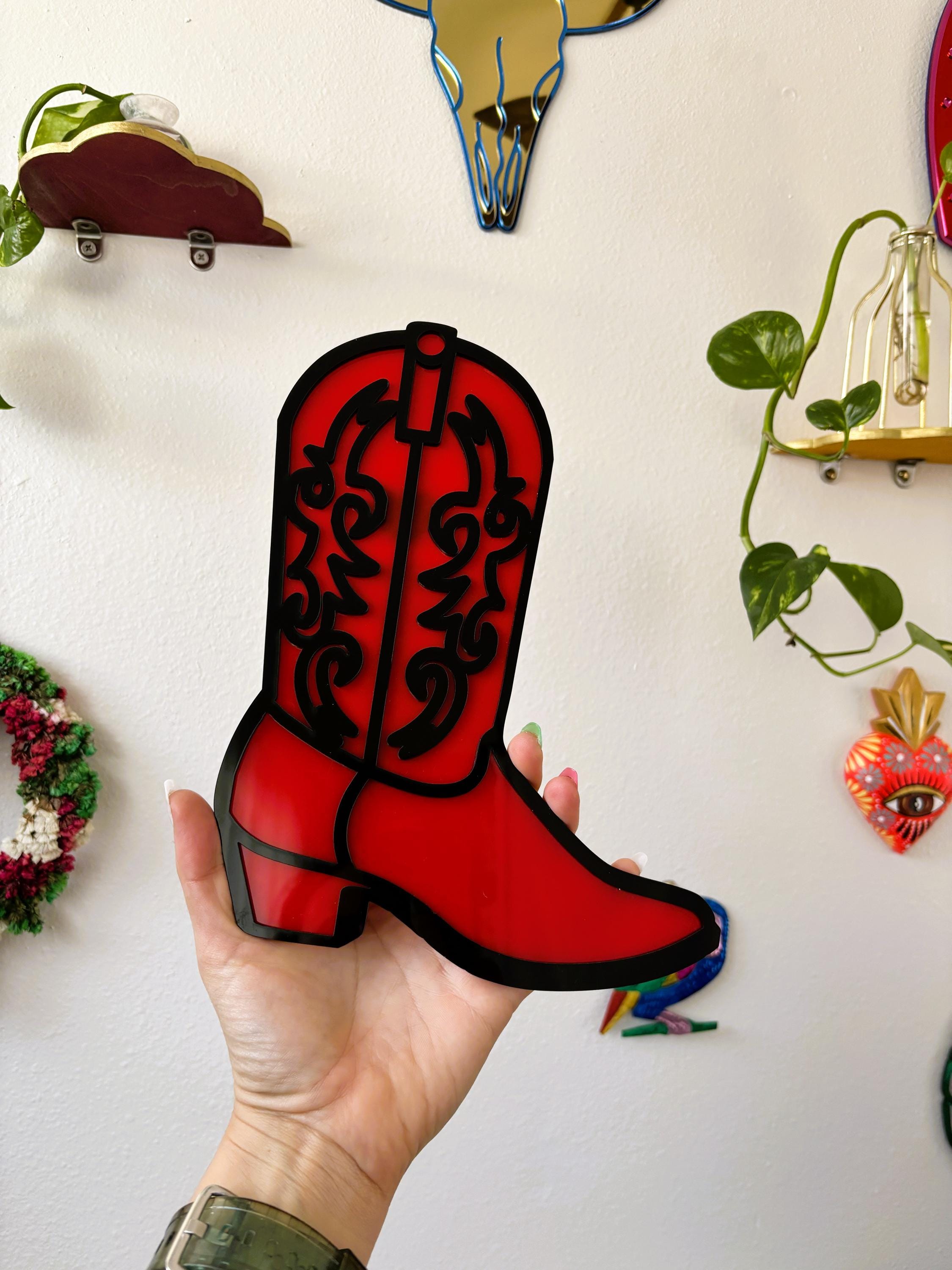 Hand holding a red cowboy boot-shaped object against a white wall with decorative items.