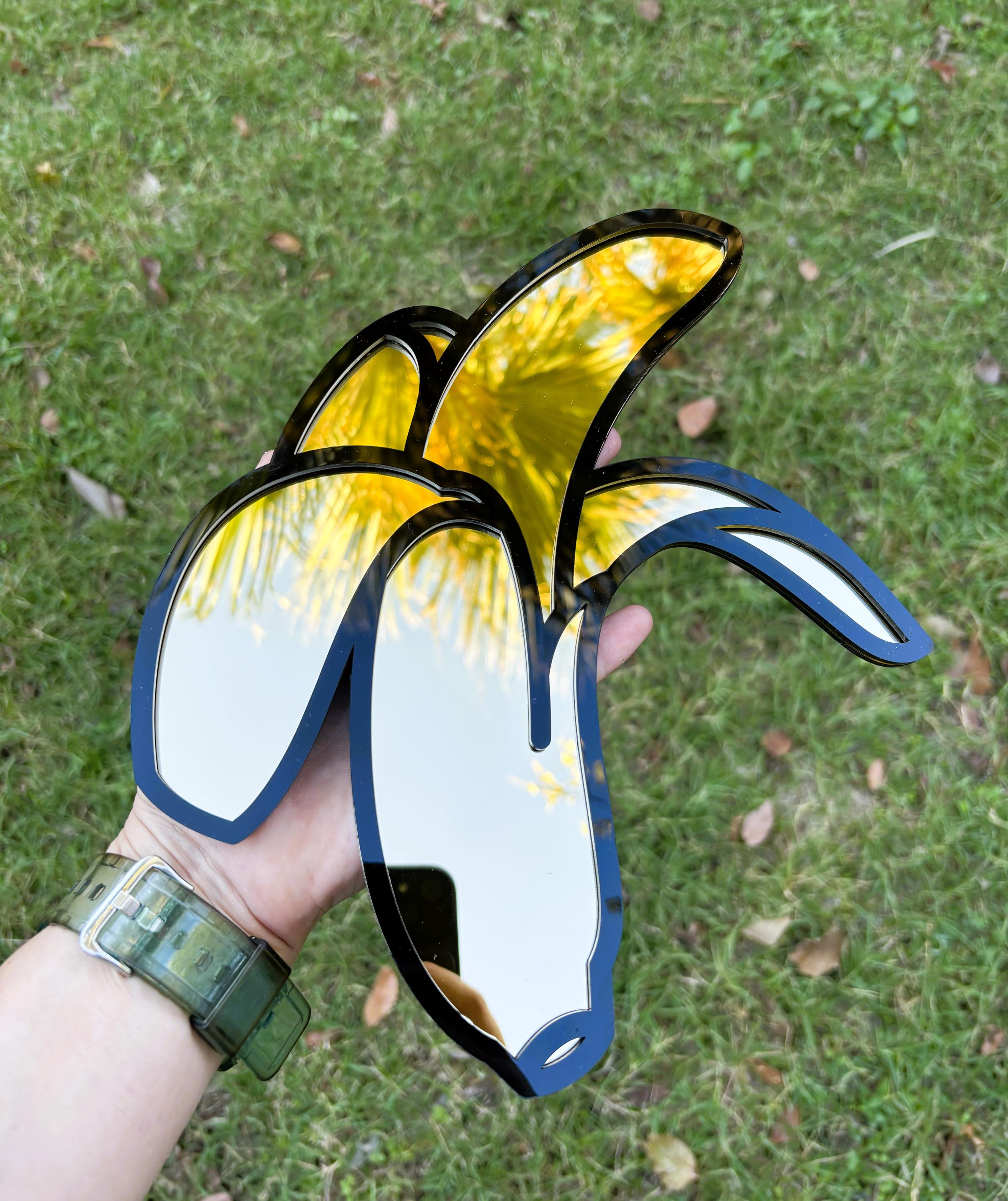 Decorative mirror shaped like a banana with yellow interior held against a grassy background.